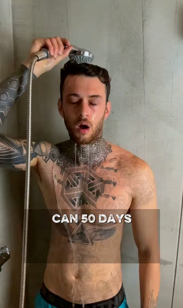 A shirtless tattooed man standing in a shower while holding a handheld shower head over his head. Text overlay reads 'CAN 50 DAYS'.