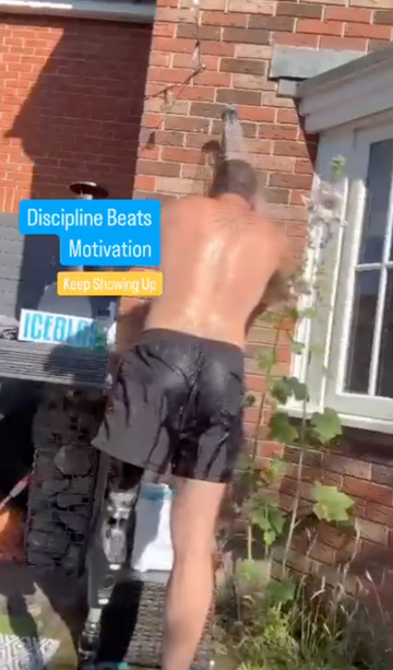 A shirtless man with a tattoo stands outdoors in front of a brick wall, wearing black swim trunks that are visibly wet. A text overlay on the left reads 'Discipline Beats Motivation' and 'Keep Showing Up'.