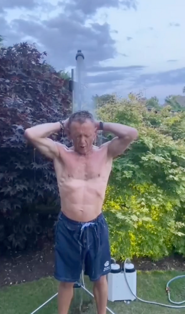 A shirtless man standing outdoors under a sprinkler in a garden, wearing dark blue swim trunks. Greenery and a cloudy sky are visible in the background.