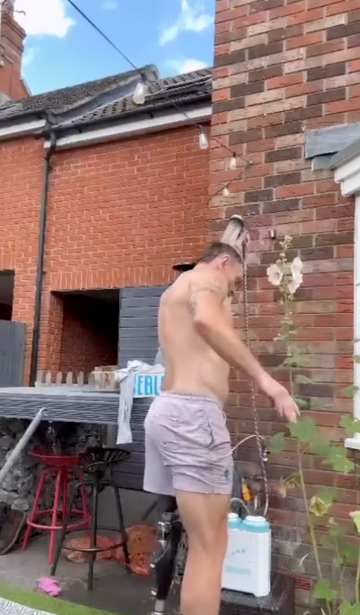 A shirtless man wearing light gray shorts stands in a backyard next to a brick wall, holding a white sprayer bottle connected to a hose. A red stool, brick walls, and outdoor plants are visible in the background.