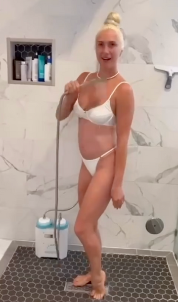 A pregnant woman with blonde hair in a bun stands in a tiled shower, holding a handheld shower head. She is wearing a white bikini.