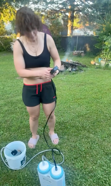 A woman standing on a grassy backyard lawn wearing a black tank top and black athletic shorts with red drawstrings, holding a phone while standing next to a white bucket and a white and blue pump device connected by a hose.