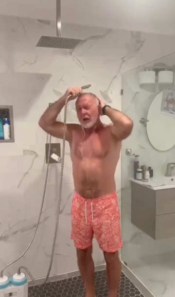 A man with white hair stands in a tiled bathroom shower, holding a handheld shower head and spraying water. He is wearing coral pink patterned swim trunks.